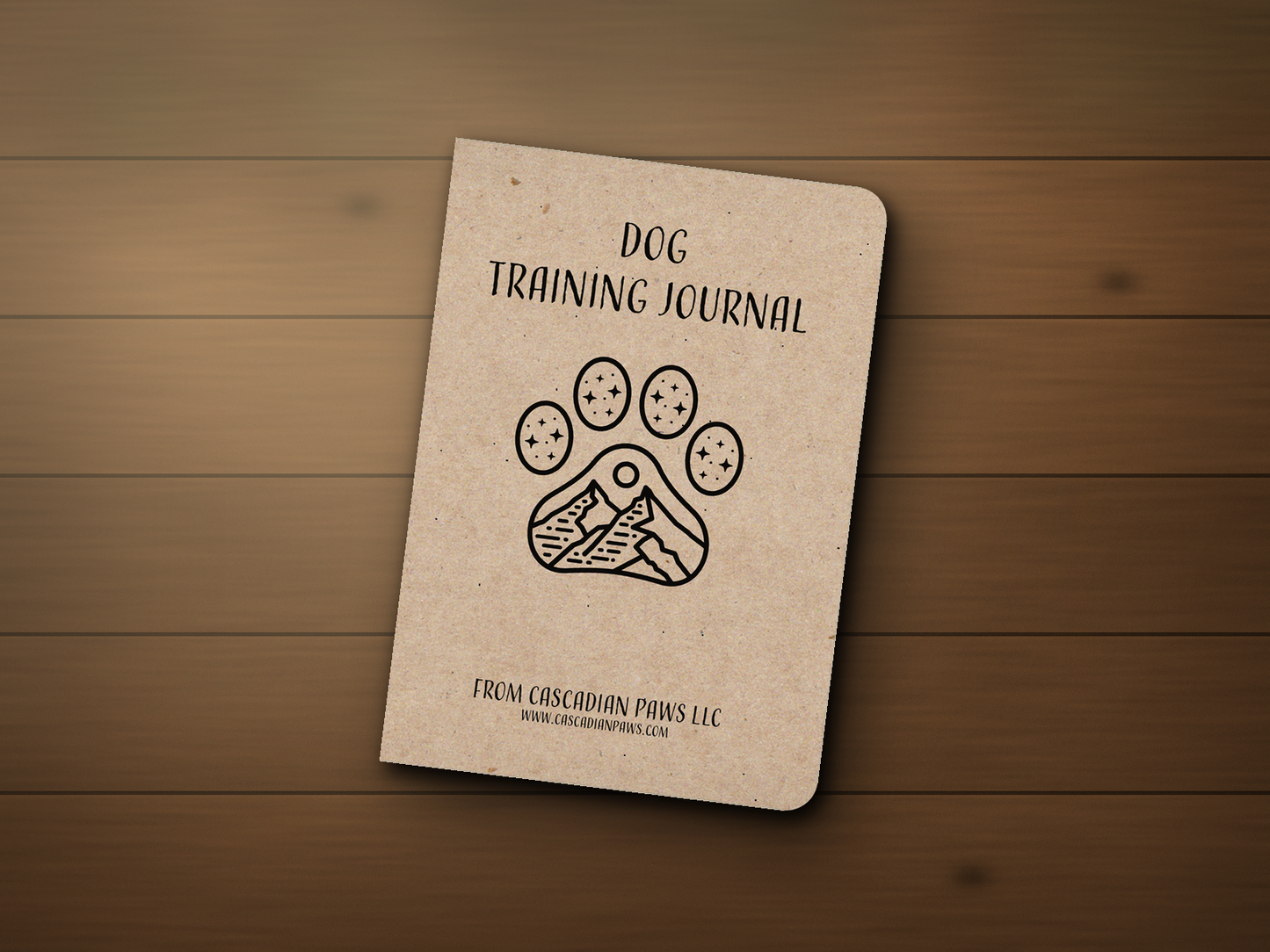Cascadian Paws Dog Training Journal — a kraft pocket notebook with a paw-and-mountain line logo, resting on a wooden outdoor patio table.