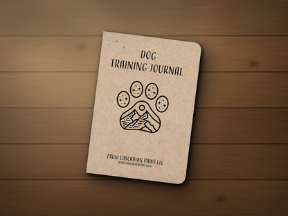 Cascadian Paws Dog Training Journal — a kraft pocket notebook with a paw-and-mountain line logo, resting on a wooden outdoor patio table.